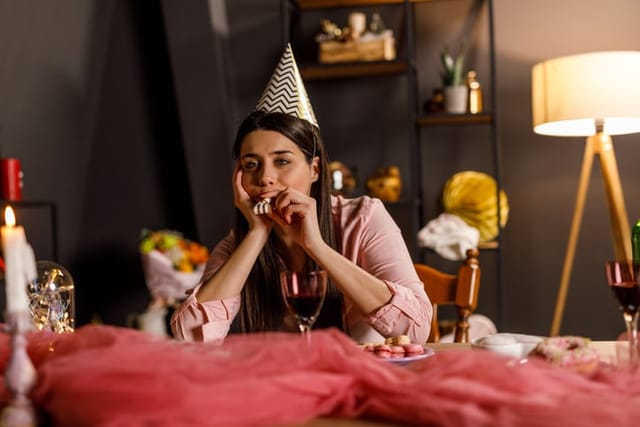 Copy space shot of bored young woman sitting at table with hand under chin, wearing a fun party hat and blowing a party horn while celebrating her birthday alone.