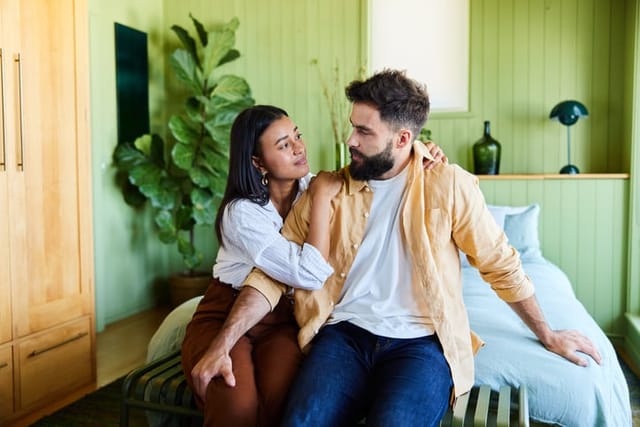 Young woman talking with her upset husband while sitting together in their bedroom at home