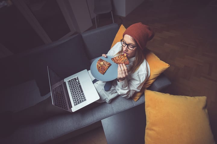 Young woman wearing shirt and a tie,lying in the bed with a laptop on her legs and eating pizza